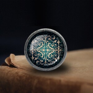 May include: A round, silver-toned knob with a decorative, teal and gold design. The design features intricate swirls and patterns.