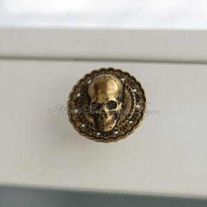 May include: A bronze cabinet knob featuring a skull design. The skull is set within a decorative, circular backing with an ornate, filigree pattern. The knob is mounted on a white surface.