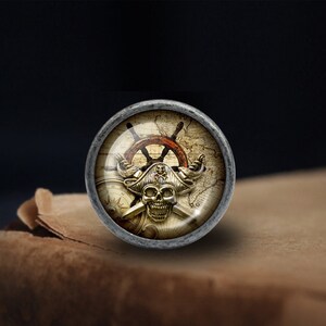 May include: A silver-toned round knob featuring a pirate skull and crossbones design. The skull is wearing a captain's hat and is surrounded by a compass, a ship's wheel, and a map.