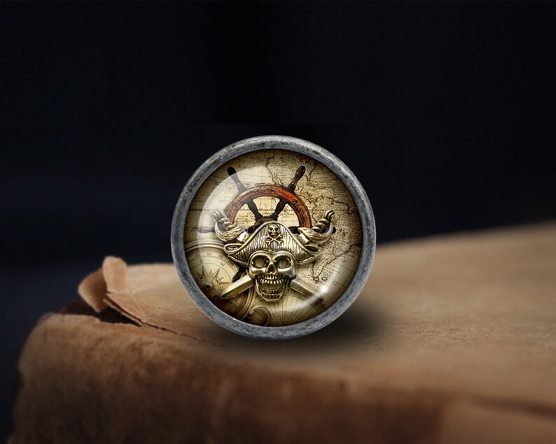 May include: A silver-toned round knob featuring a pirate skull and crossbones design. The skull is wearing a captain's hat and is surrounded by a compass, a ship's wheel, and a map.