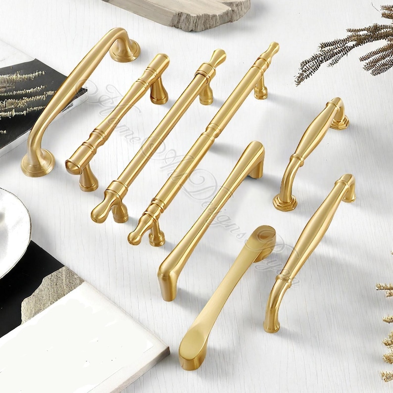 May include: A collection of gold-colored cabinet pulls and handles in various designs. The hardware includes both curved and straight pulls, with different decorative details. The items are arranged on a white surface, showcasing their metallic finish.