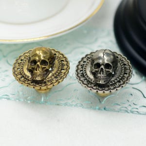 May include: Two decorative cabinet knobs with skull designs. One is gold-toned, the other silver-toned. Each knob features a skull in the centre, surrounded by an ornate, circular border. The knobs are likely made of metal.