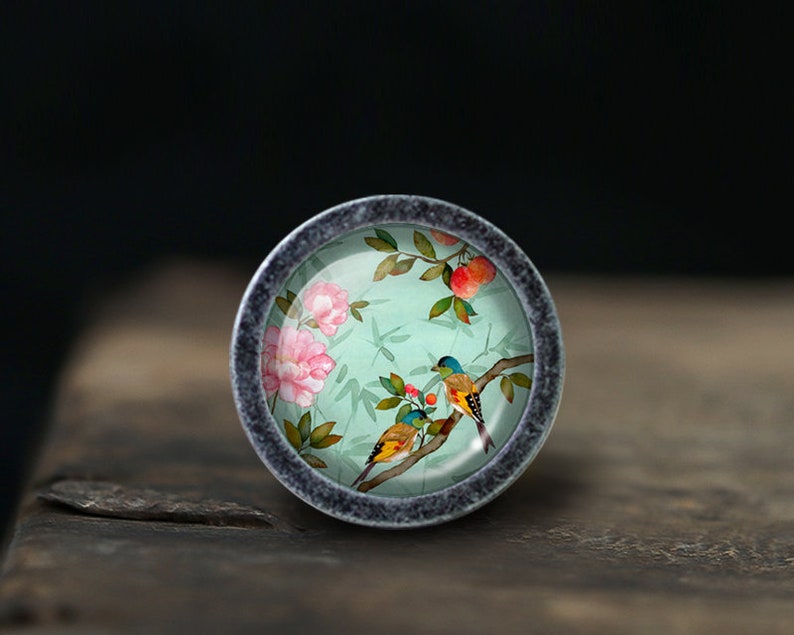 May include: A round, silver-toned cabinet knob with a floral design. The knob features a light blue background with pink flowers, green leaves, and yellow and blue birds.