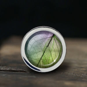 Może przedstawiać: Srebrny pierścionek z przezroczystą kopułą z żywicy, w której znajduje się sprasowany liść w odcieniach zieleni, fioletu i bieli.