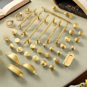 Puede incluir: Una colecci&oacute;n de herrajes dorados para gabinetes, que incluyen perillas, tiradores y manijas. Los herrajes presentan varios dise&ntilde;os, desde perillas redondas simples hasta tiradores ornamentados con detalles decorativos.