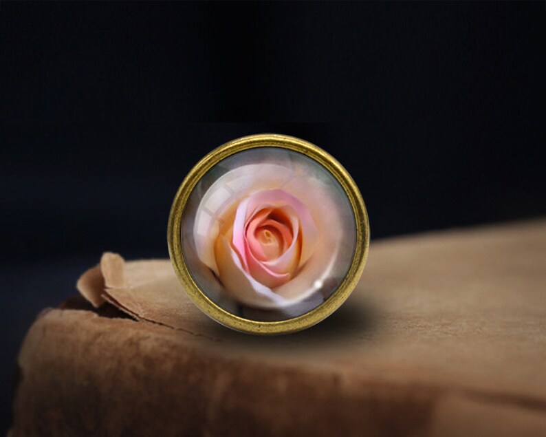 Op de afbeelding: Een ronde knop in goudkleur met een glazen koepel met een roze en witte roos.