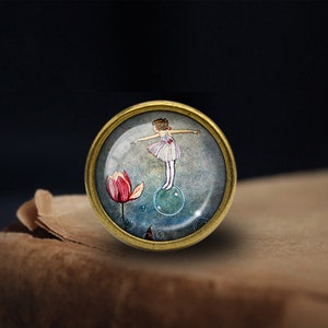 May include: A round glass cabochon with a gold metal frame features a watercolor illustration of a girl in a white dress standing on a bubble with a pink flower in the background.