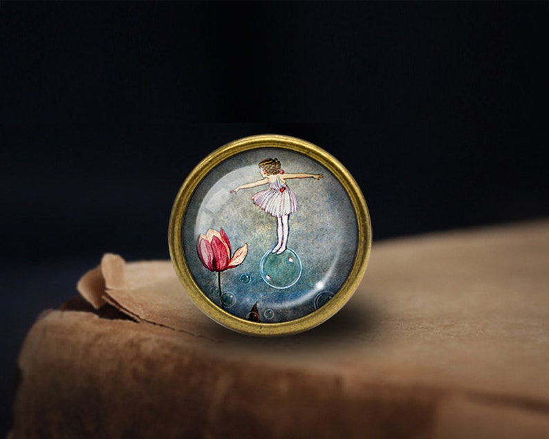 May include: A round glass cabochon with a gold metal frame features a watercolor illustration of a girl in a white dress standing on a bubble with a pink flower in the background.