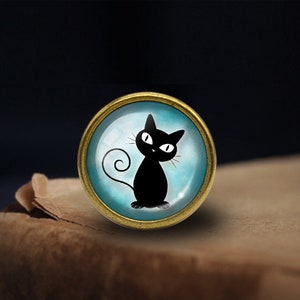 Peut inclure: Une silhouette de chat noir avec des yeux blancs sur un fond bleu, encadr&eacute;e dans un cercle m&eacute;tallique dor&eacute;.