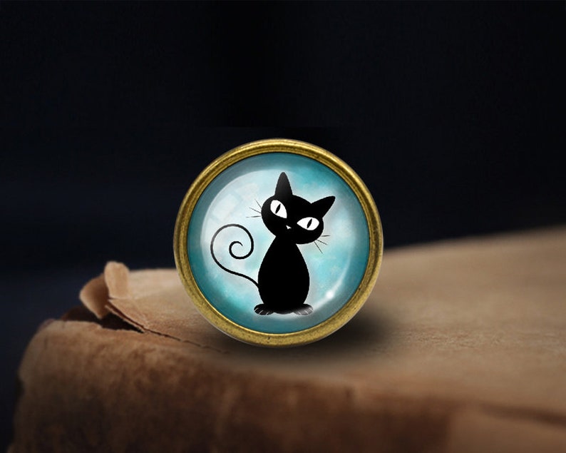 Peut inclure: Une silhouette de chat noir avec des yeux blancs sur un fond bleu, encadr&eacute;e dans un cercle m&eacute;tallique dor&eacute;.