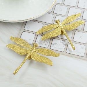 May include: Two gold-toned dragonfly drawer pulls with a textured finish. The pulls are shown on a white surface.