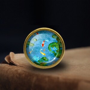 Peut inclure: Une &eacute;pingle ronde en m&eacute;tal dor&eacute; avec un d&ocirc;me en verre repr&eacute;sentant une peinture &agrave; l'aquarelle bleue et verte de poissons ko&iuml; nageant dans un &eacute;tang avec des n&eacute;nuphars.