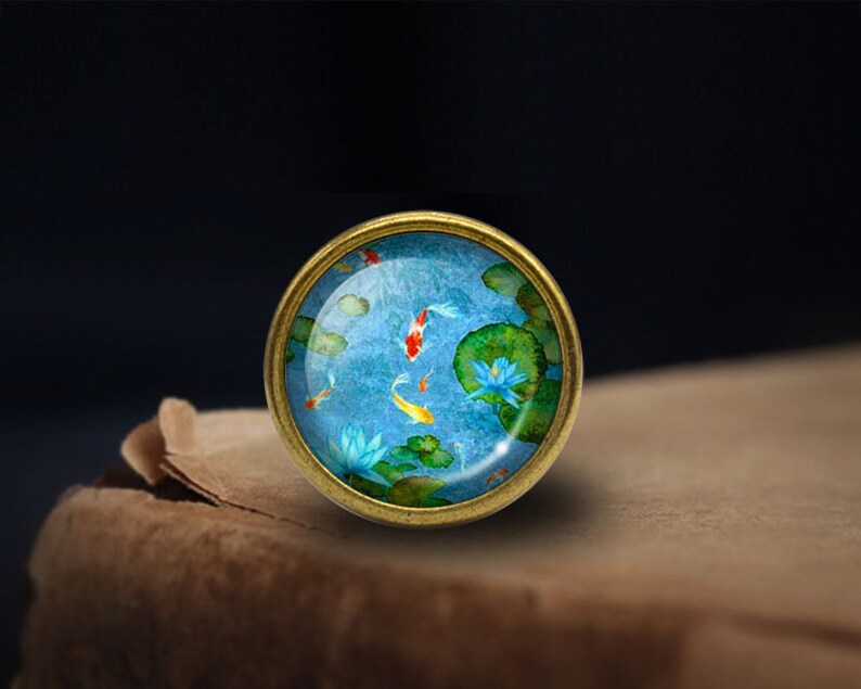 Peut inclure: Une &eacute;pingle ronde en m&eacute;tal dor&eacute; avec un d&ocirc;me en verre repr&eacute;sentant une peinture &agrave; l'aquarelle bleue et verte de poissons ko&iuml; nageant dans un &eacute;tang avec des n&eacute;nuphars.