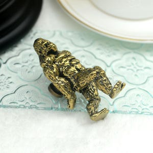 K&ouml;nnte beinhalten: Ein goldfarbener Metall-Schrankknauf in Form eines sitzenden Gorillas. Der Gorilla ist mit strukturiertem Fell detailliert und hat eine entspannte Pose.