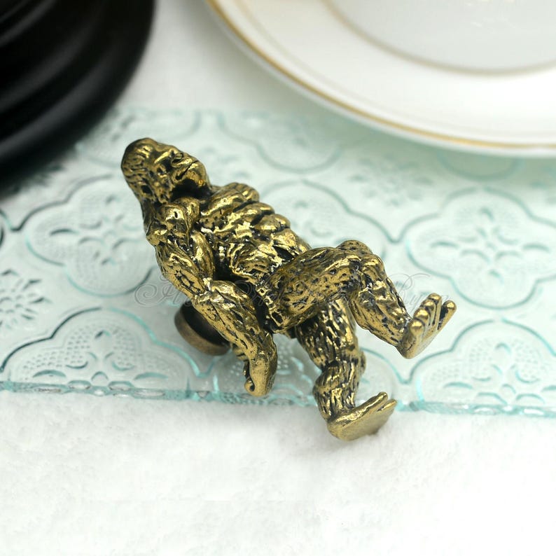 K&ouml;nnte beinhalten: Ein goldfarbener Metall-Schrankknauf in Form eines sitzenden Gorillas. Der Gorilla ist mit strukturiertem Fell detailliert und hat eine entspannte Pose.