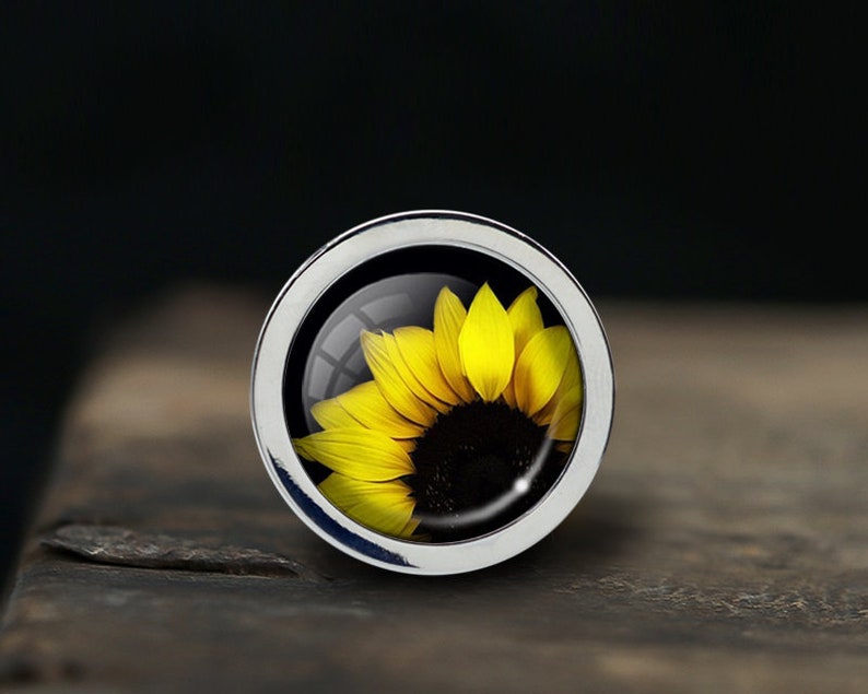 Op de afbeelding: Een zilveren ring met een glazen koepel die een gele zonnebloem toont.