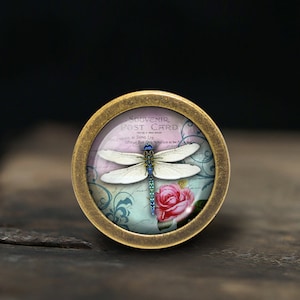 Puede incluir: Un colgante redondo de color latón con una cúpula de vidrio que presenta un diseño de postal vintage. La postal representa una libélula con alas azules y una rosa rosa. El texto de la postal dice "Souvenir Post Card".