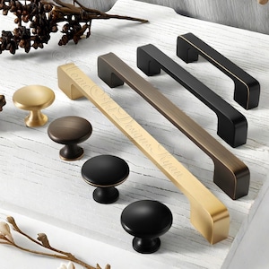 May include: A selection of cabinet knobs and pulls in gold, bronze, and black finishes. The image shows a variety of designs, including round knobs and rectangular pulls. One pull has the text "Home Art Designs Ryan".