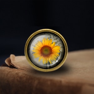 May include: A round, gold-toned metal pin with a glass dome featuring a yellow sunflower with a brown center. The sunflower is surrounded by a white, textured background.