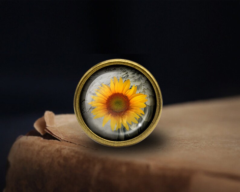 May include: A round, gold-toned metal pin with a glass dome featuring a yellow sunflower with a brown center. The sunflower is surrounded by a white, textured background.
