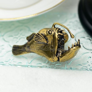 K&ouml;nnte beinhalten: Bronze farbene Anglerfisch-Figur mit offenem Maul, das scharfe Z&auml;hne zeigt. Der Fisch hat detaillierte Flossen und ein gl&uuml;hbirnenartiges Anh&auml;ngsel, das sich von seinem Kopf erstreckt. Die Figur wird auf einer strukturierten Glasoberfl&auml;che pr&auml;sentiert.