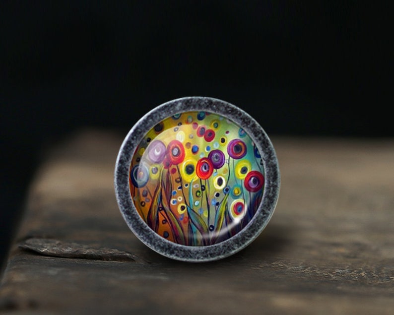 May include: A round, silver-toned knob with a colorful abstract floral design. The design features a field of flowers with yellow, red, blue, and purple petals. The knob is perfect for adding a touch of whimsy to any room.
