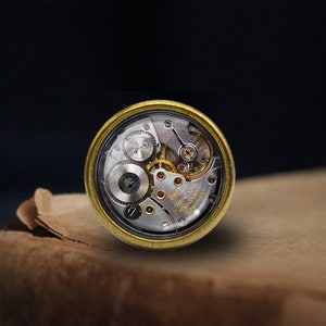 May include: A gold-toned watch movement with a clear glass cover, revealing the intricate inner workings of the mechanism. The watch movement is labelled "17 Jewels 12.50mm Swiss Made" and "Longines".