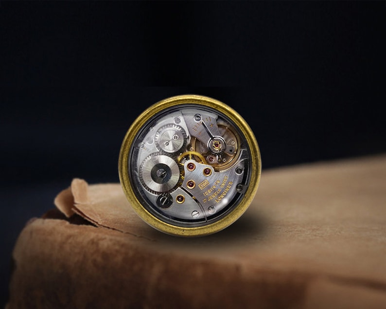 May include: A gold-toned watch movement with a clear glass cover, revealing the intricate inner workings of the mechanism. The watch movement is labelled "17 Jewels 12.50mm Swiss Made" and "Longines".