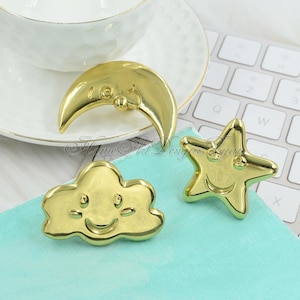 Può includere: Tre pomelli decorativi dorati a forma di luna, nuvola e stella sorridenti. I pomelli sono su una superficie azzurra, con una tazza da tè bianca e una tastiera sullo sfondo.