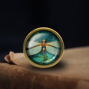 May include: A gold-toned metal ring with a glass cabochon featuring a detailed dragonfly design. The dragonfly has a white body with gold accents and is set against a dark blue background with a starry effect.