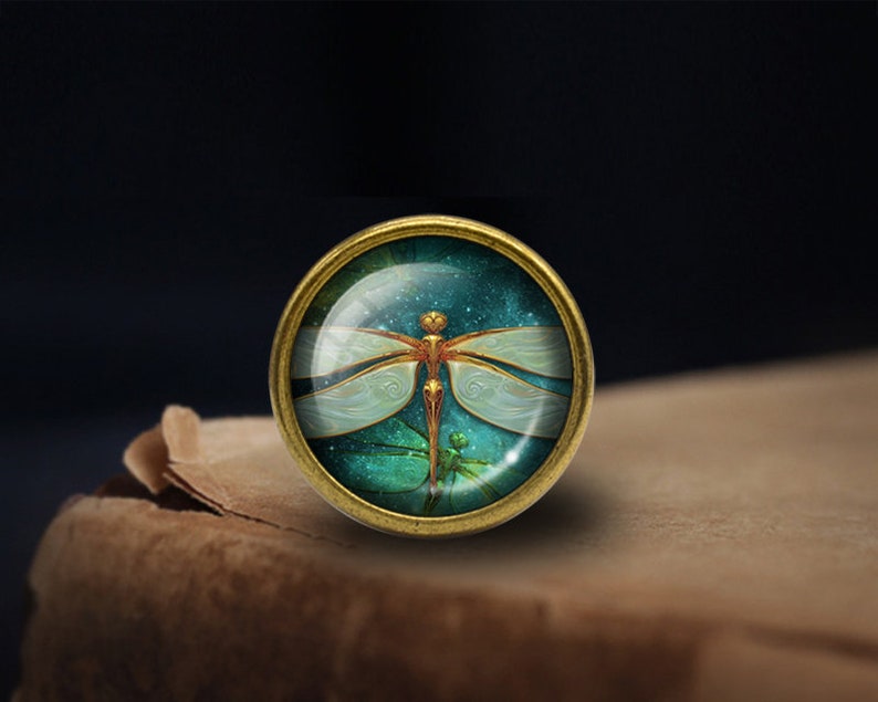 May include: A gold-toned metal ring with a glass cabochon featuring a detailed dragonfly design. The dragonfly has a white body with gold accents and is set against a dark blue background with a starry effect.
