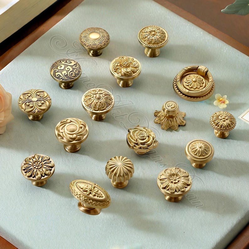 May include: A selection of antique brass cabinet knobs featuring diverse floral and geometric patterns. The knobs vary in shape and size, including round, oval, and flower-shaped, with detailed ornamentation. The knobs are presented on a light blue surface.