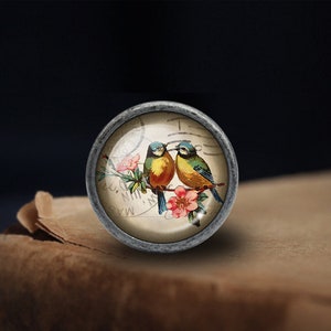 May include: A vintage-style glass cabochon with a silver-toned metal setting features a pair of bluebirds perched on a branch with pink flowers. The birds are yellow and blue with black eyes. The background is a vintage-style postcard with the text "MAY. N.Y.  N.M."