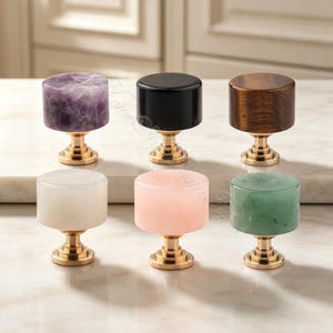 Puede incluir: Un conjunto de seis pomos cilíndricos para armarios dispuestos en dos filas. Los pomos presentan diversos materiales, incluyendo amatista morada, negro sólido, madera marrón, piedra blanca, rosa claro y verde, montados sobre bases de latón.