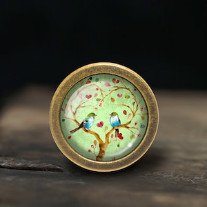 Può includere: Un pomello rotondo color ottone con un inserto in vetro verde che presenta un disegno dipinto di due uccelli blu appollaiati su un ramo d'albero con cuori rosa e foglie verdi.