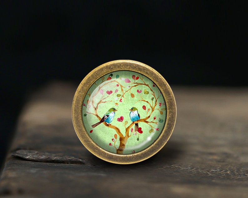 Puede incluir: Un pomo redondo de color lat&oacute;n con un inserto de vidrio verde que presenta un dise&ntilde;o pintado de dos p&aacute;jaros azules posados en una rama de &aacute;rbol con corazones rosas y hojas verdes.