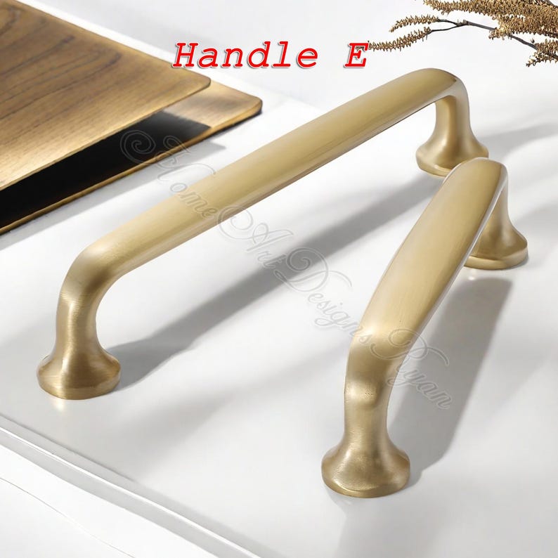 Puede incluir: Dos tiradores de armario de color lat&oacute;n. Uno es largo y rectangular, y el otro es m&aacute;s corto. Los tiradores tienen un dise&ntilde;o curvo y est&aacute;n colocados sobre una superficie blanca. El texto "Handle E" es visible.