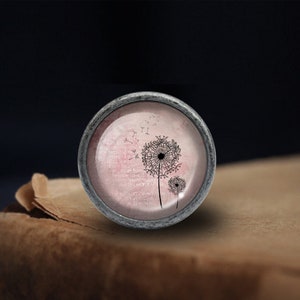 Peut inclure: Un bouton rond argent&eacute; avec un fond rose et blanc repr&eacute;sentant une silhouette noire de pissenlit avec des graines qui s'envolent dans le vent.