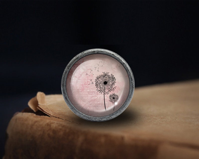 Peut inclure: Un bouton rond argent&eacute; avec un fond rose et blanc repr&eacute;sentant une silhouette noire de pissenlit avec des graines qui s'envolent dans le vent.