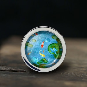 Könnte beinhalten: Ein silberner Ring mit einem blauen und grünen Emaille-Design von Koi-Karpfen, die in einem Teich mit Seerosen schwimmen.