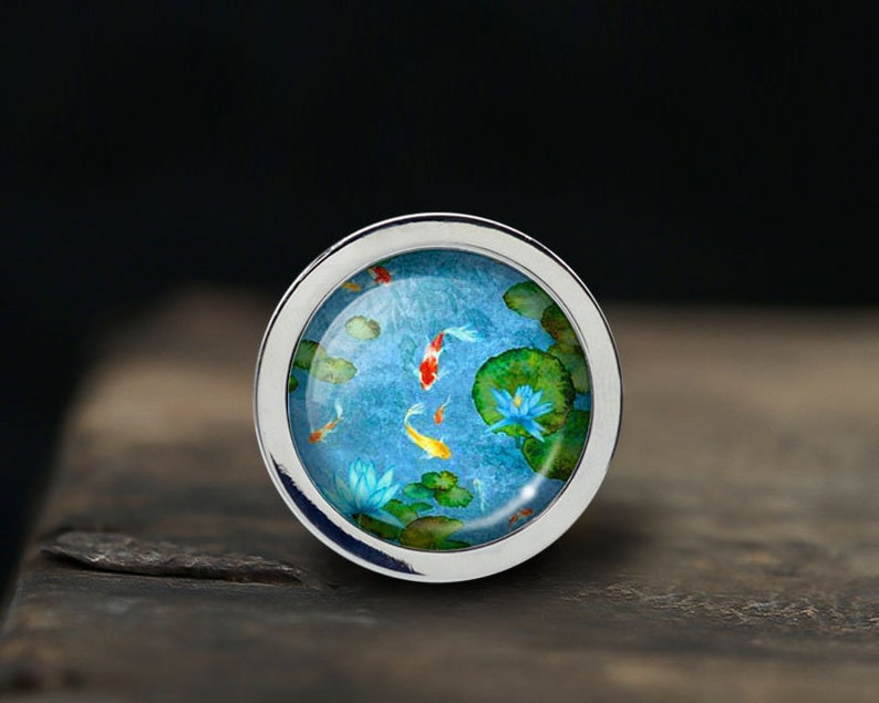 Peut inclure: Une bague en argent avec un motif en &eacute;mail bleu et vert repr&eacute;sentant des carpes ko&iuml; nageant dans un &eacute;tang avec des n&eacute;nuphars.