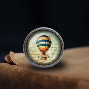K&ouml;nnte beinhalten: Ein Knopf im Vintage-Stil, silberfarben, mit einer Glasoberfl&auml;che, die eine farbenfrohe Illustration eines Hei&szlig;luftballons zeigt. Der Ballon ist gelb, blau und rot mit wei&szlig;en Streifen. Der Hintergrund ist hellblau mit handschriftlichem Text.