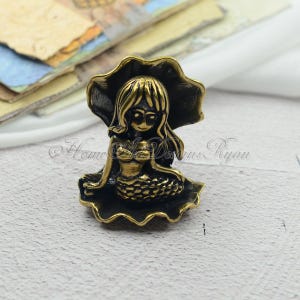 May include: A gold-toned mermaid figurine sitting on a seashell. The mermaid has long hair and is wearing a seashell bra.