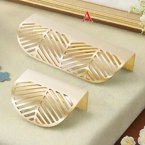 K&ouml;nnte beinhalten: Zwei dekorative Schrankgriffe mit Blattdesign. Die Griffe sind cremefarben mit goldenen Akzenten und einem ausgeschnittenen Bl&auml;ttermuster. Die Griffe haben eine gebogene Form und sind aus Metall.