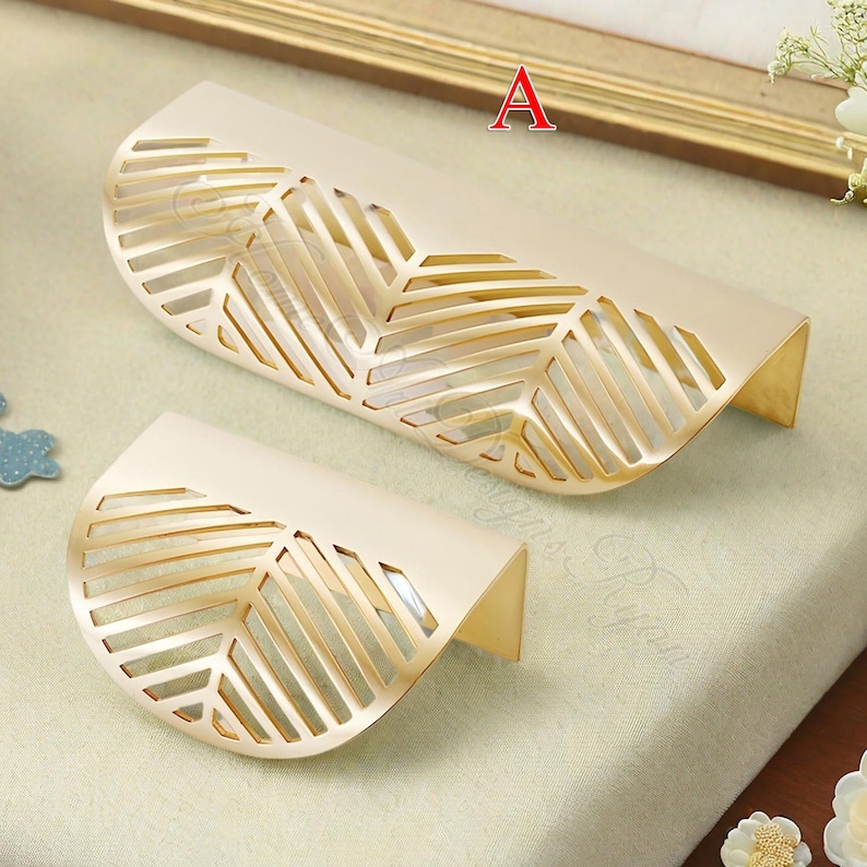 K&ouml;nnte beinhalten: Zwei dekorative Schrankgriffe mit Blattdesign. Die Griffe sind cremefarben mit goldenen Akzenten und einem ausgeschnittenen Bl&auml;ttermuster. Die Griffe haben eine gebogene Form und sind aus Metall.