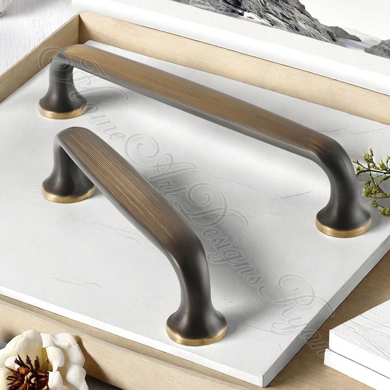K&ouml;nnte beinhalten: Zwei bronzefarbene Schrankgriffe mit geschwungenem Design. Die Griffe haben eine dunkle Basis mit einem goldfarbenen Akzent am Boden. Die Griffe sind auf einer wei&szlig;en Oberfl&auml;che platziert.
