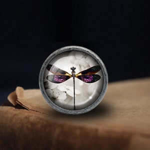 May include: A silver-toned round knob with a glass insert featuring a purple and yellow dragonfly on a white background.