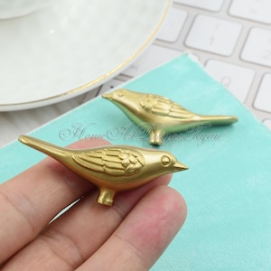 May include: Two gold bird-shaped drawer pulls with a detailed design. The pulls are made of metal and have a shiny finish.