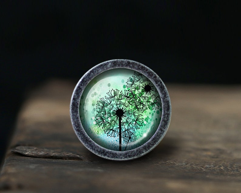 May include: A silver-toned round button with a glass top featuring a watercolour-style design of three dandelion flowers in shades of green and black.