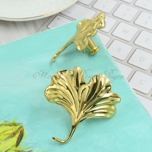 Könnte beinhalten: Zwei goldfarbene Haken in Form von Ginkgoblättern. Die Haken befinden sich auf einer türkisfarbenen Oberfläche.